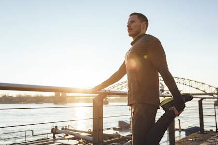 Sportler steht an einem Geländer und dehnt seine Oberschenkelvorderseite.