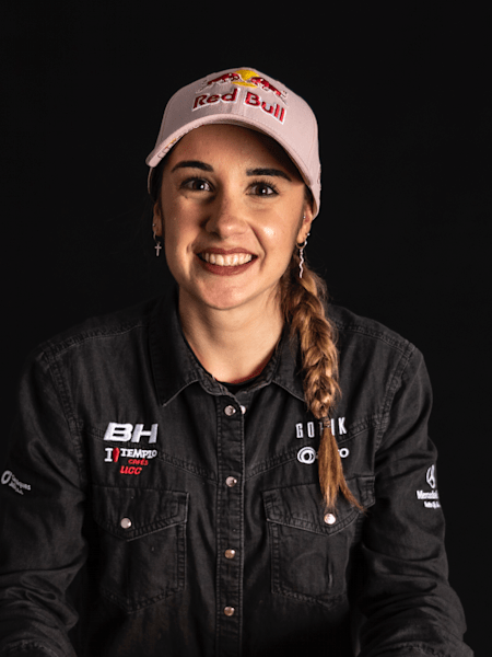Portrait de la piloto de MTB XCO Rocío del Alba, sonriendo, con gorra Red Bull de color rosa, una trenza y una camisa negra con el nombre de sus sponsors en blanco. El fondo es negro.