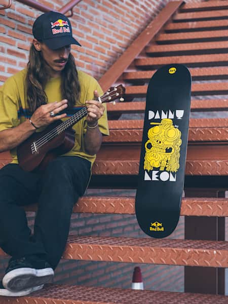 El skater mostoleño Danny León tocando el ukelele al lado de la tabla de skate que le ha pintado el artista urbano Juan Díaz-Faes con un retrato suyo y el clavijero del ukelele asomando.
