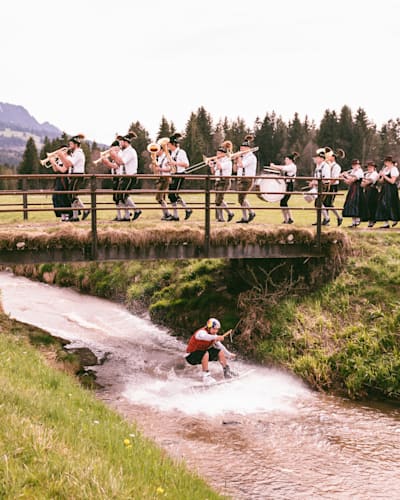 Wakeboard-Profi Felix Georgii gleitet auf einem Fluss unter einer Brücke durch, auf der eine Blaskappelle spielt.