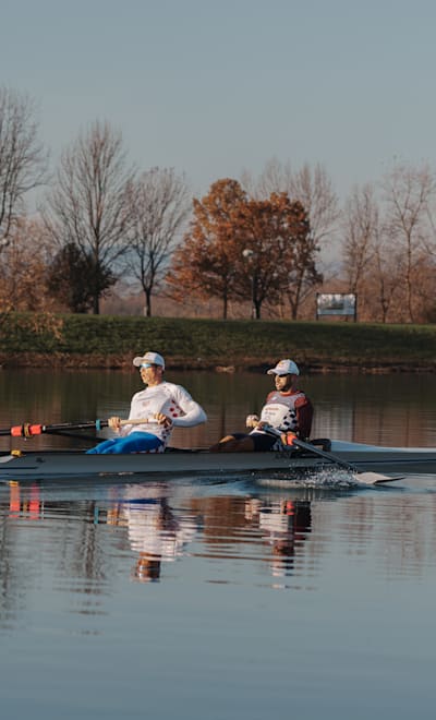 Valent Sinković and Martin Sinković rowing on a tranquil Jarun Lake in Zagreb, Croatia, surrounded by autumn foliage.
