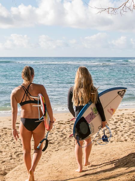 Les surfeuses Carissa Moore et Caroline Marks marchent sur la plage avec leurs planches de surf. Retour sur dix surfeuses qui ont marqué l'histoire du surf.