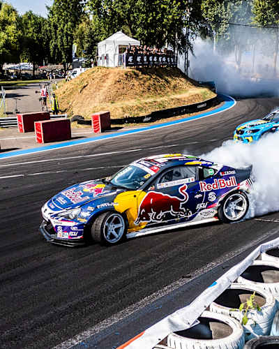 Two cars are in a controlled slide, kicking up a cloud of tyre smoke as they race in a thrilling competition called Drift Masters, where both precision and speed are the key to winning.