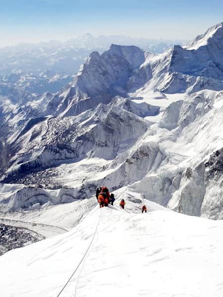 Climbers ascend on snowy peak with a mountain range in the background.