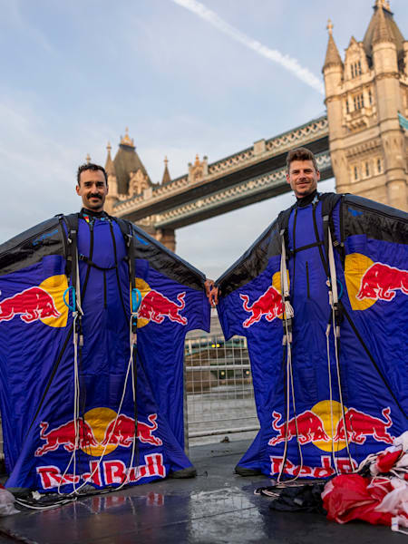 Marco Fürst și Marco Waltenspiel, în costumele lor de zbor Red Bull, în fața Tower Bridge din Londra, cu parașutele așezate pe jos lângă ei
