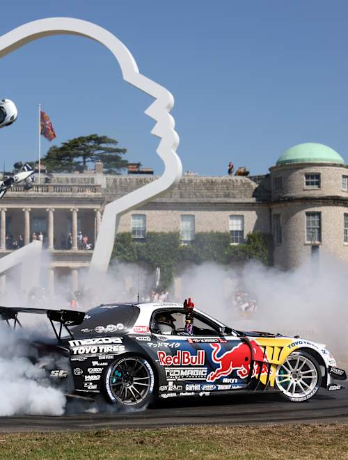 A Red Bull car drifts around Goodwood House at the 2025 Festival of Speed in the UK, with the driver waving to spectators