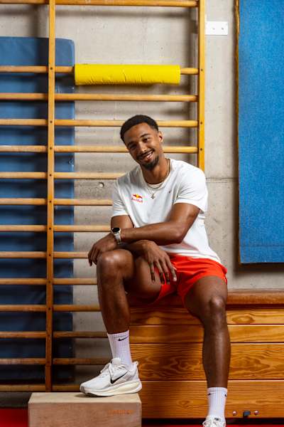 Leo Neugebauer, con la equipación de atletismo de la marca Red Bull, se sienta y sonríe en un gimnasio durante una sesión fotográfica en Alemania en 2026.