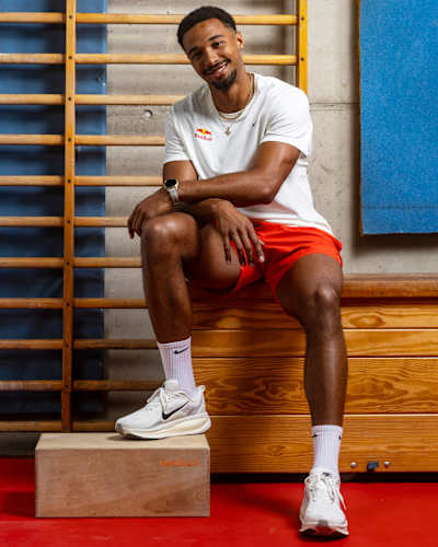Leo Neugebauer, con la equipación de atletismo de la marca Red Bull, se sienta y sonríe en un gimnasio durante una sesión fotográfica en Alemania en 2026.