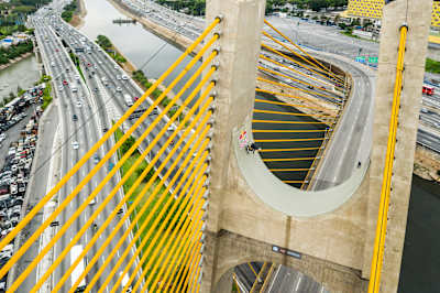 Sandro Dias skatet eine natürliche Halfpipe, hoch oben zwischen den Pfeilern einer Autobahnbrücke. Unter ihm sind Highways zu sehen und ein Fluss.