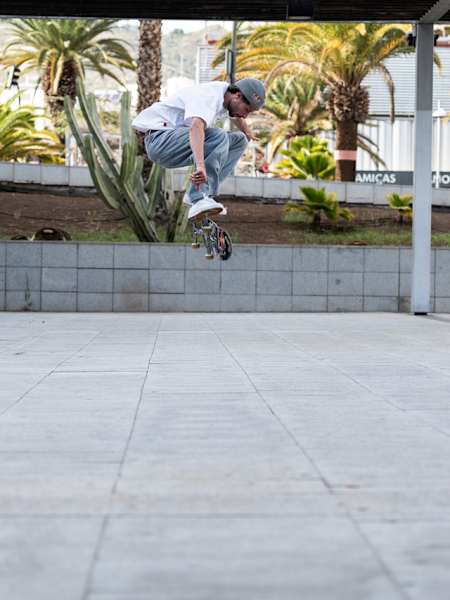 Jost Arens mit einem Kickflip auf einem offenen Platz, von vorne fotografiert. Im Hintergrund sind Palmen zu sehen.