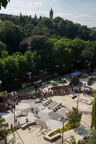 A view from above of the skate park, located in the valley, surrounded by trees and well frequented by spectators. 