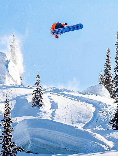 A snowboarder is in mid-air after jumping off a ramp, with snow spraying behind them against a clear sky at the Finals of Natural Selection Snowboard 2026 in Revelstoke, BC, Canada.