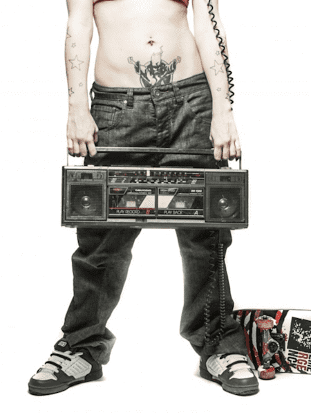 Una chica tatuada en top y pantalonas y bambas anchas sostiene un radio cassette para reproducir música al lado de una tabla de skate tirada en el suelo.
