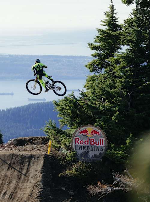 Canadian Jackson Goldstone soars over a dirt jump at Red Bull Hardline in British Columbia, Canada, with forested mountains and a lake behind him on October 15 2026