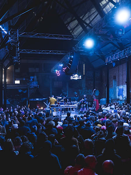 Une foule vibre devant un DJ lors d’un festival de musique électronique, sous une verrière industrielle illuminée. 