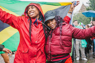 Deux festivalières sourient sous la pluie devant un drapeau jamaïcain lors d’un événement sound system à Londres.
