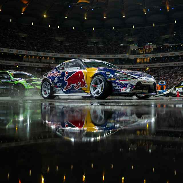 Two cars are in a controlled slide, their reflections visible on the slick track, as they race in a thrilling competition called Drift Masters, where both precision and speed are the key to winning.