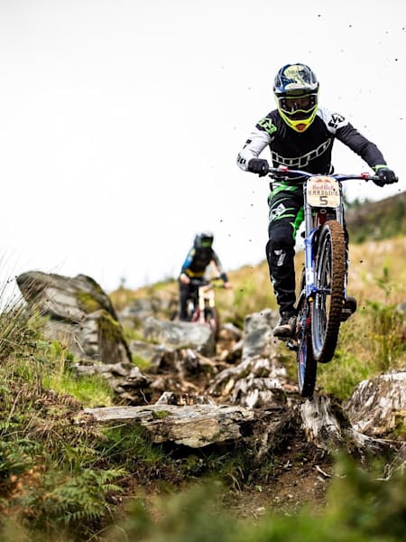 Adam Brayton rides down the Red Bull Hardline track while being followed by another rider.