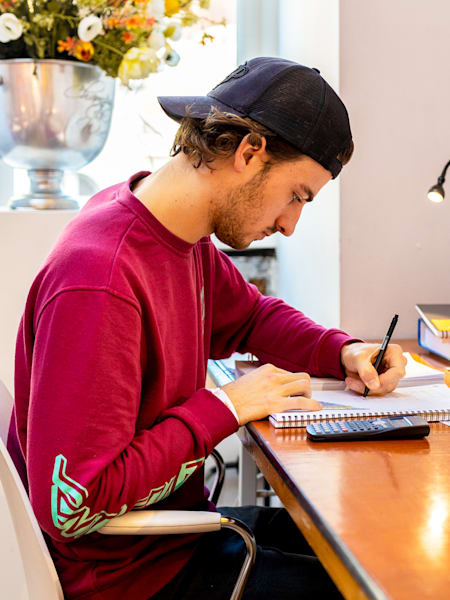 Student siedzący przy biurku i robiący notatki, obok niego leży kalkulator i stoi kubek z Red Bullem.