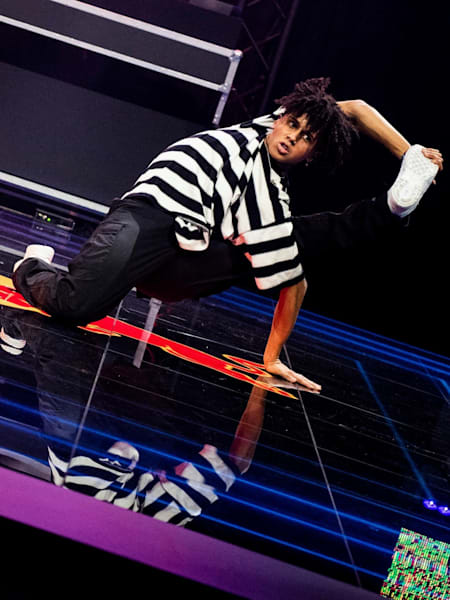 Shinshan is on the floor and holds one leg with a hand that reaches behind his head.