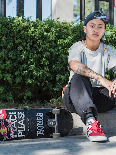 Dziewczyna w szarej koszulce siedzi na krawężniku, a po jej prawej stronie leży czarna deskorolka.