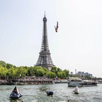A view from the Seine of the Eiffel Tower with a diver in mid-air in front of it. 