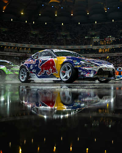 Two cars are in a controlled slide, their reflections visible on the slick track, as they race in a thrilling competition called Drift Masters, where both precision and speed are the key to winning.