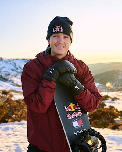 In 2023, Scotty James smiles while holding a snowboard at Perisher Resort, Australia against a scenic snowy mountain backdrop.