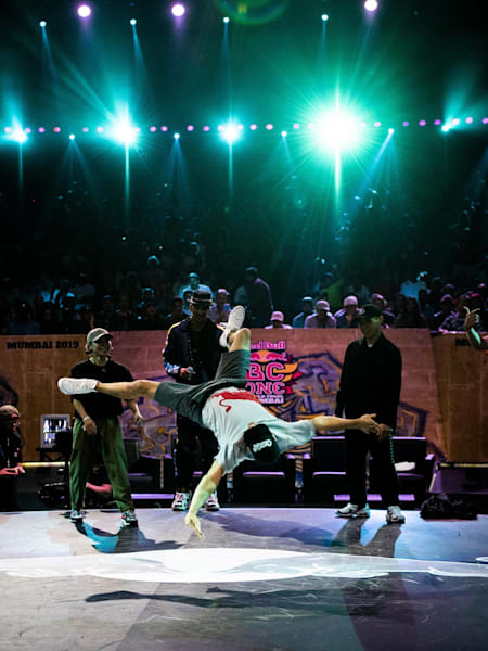 Lil G does airflares on the stage of BC One 2019. The other judges are standing in the background.