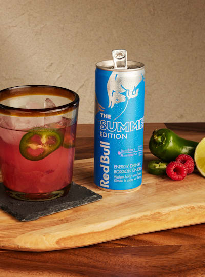 A spicy raspberry punch mocktail in a glass, an open can of Red Bull Summer Edition Juneberry, and a jalapeno pepper, lemon slice and two raspberries.