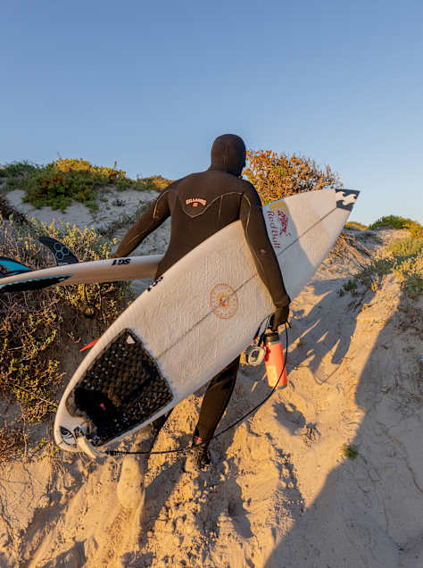 A surfer carries two boards under arm toward the waves in South Africa, for Natural Selection Tour Surf 2026