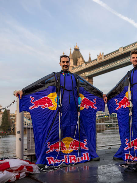 Marco Fürst und Marco Waltenspiel stehen in ihren Red Bull Wingsuits vor der Tower Bridge in London, die Fallschirme liegen neben ihnen auf dem Boden