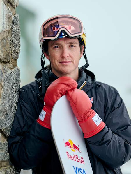 In August 2025, Scotty James stands ready at Perisher Village, Australia,  gripping his snowboard and leaning against a stone wall.