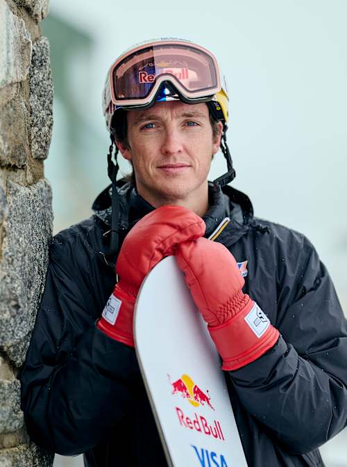 In August 2025, Scotty James stands ready at Perisher Village, Australia,  gripping his snowboard and leaning against a stone wall.