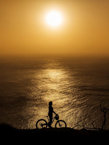 A mountain biker admires a sunset from a high vantage point  on an ocean-top cliff.