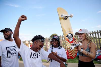 Leticia Bufoni hält auf einer Veranstaltung in Johannesburg ein Skateboard in die Luft, neben ihre stehen zwei Frauen und ein Mann, beide Frauen lachen, eine tanzt.