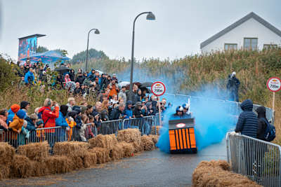 A creative soapbox car speeds through blue smoke as crowds cheer during Red Bull Olabil 2025 in Trondheim, Norway, capturing the excitement and energy of this iconic Red Bull event
