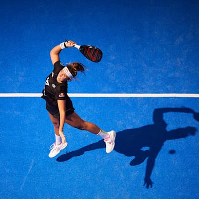 Bird's-eye view of Bea Gonzalez taking a swing as the ball approaches, silhouette reflected on blue court, at the Sevilla Premier Padel P2 final in Sevilla, Spain, 2024