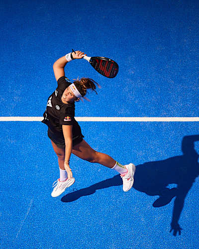 Bird's-eye view of Bea Gonzalez taking a swing as the ball approaches, silhouette reflected on blue court, at the Sevilla Premier Padel P2 final in Sevilla, Spain, 2024