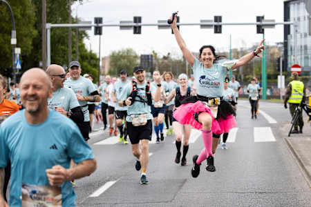 Runner in black and pink tutu jubilantly jumping during the Wings for Life World Run in Poznan, Poland amongst a crowd of other runners.