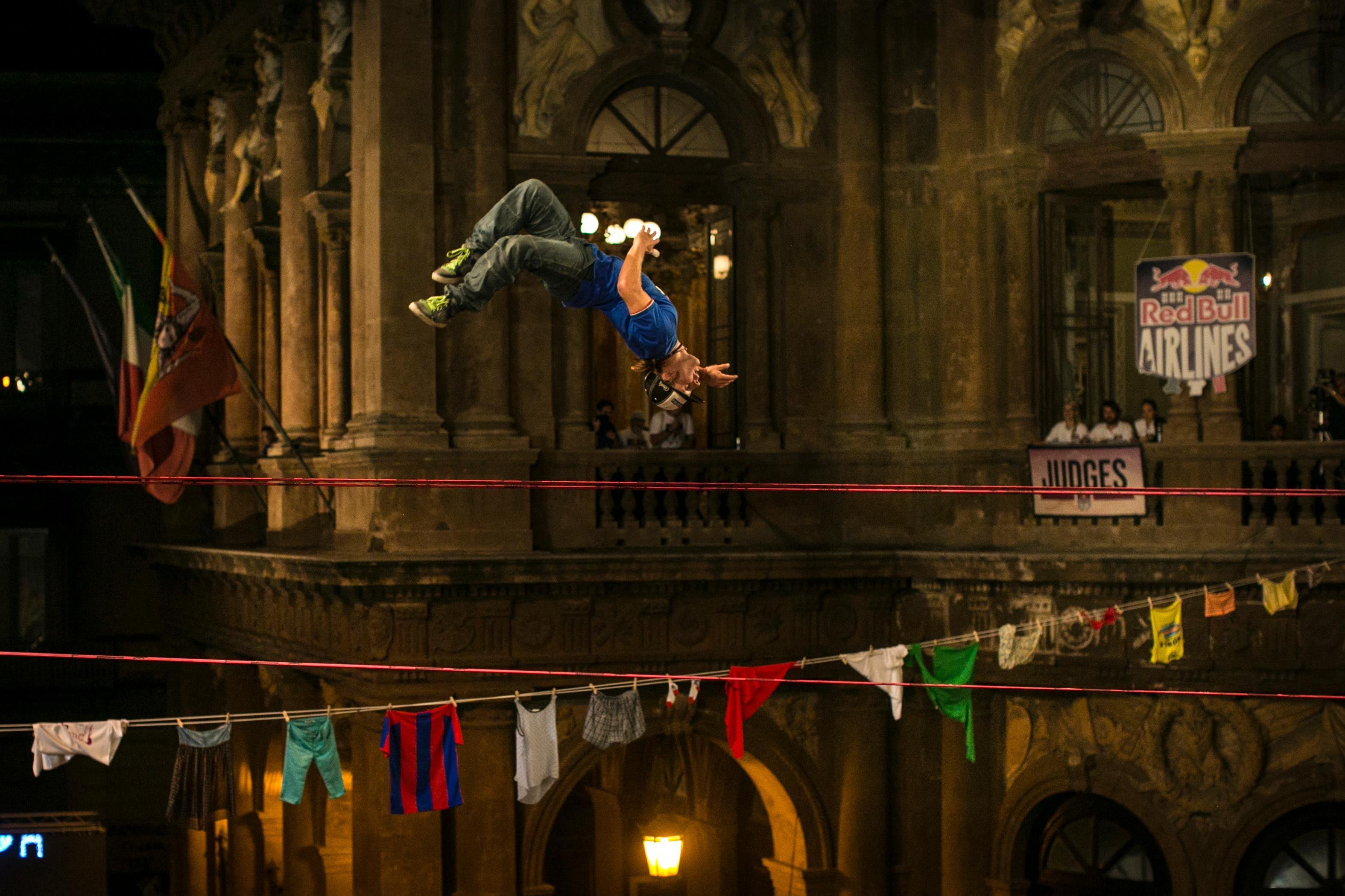 A slackliner performs a backflip on a trickline stretched across an ornate building facade at night. Below the line, colorful clothing items are hung. A Red Bull Airlines banner and judges' area are visible in the background. Slackline סלקליין